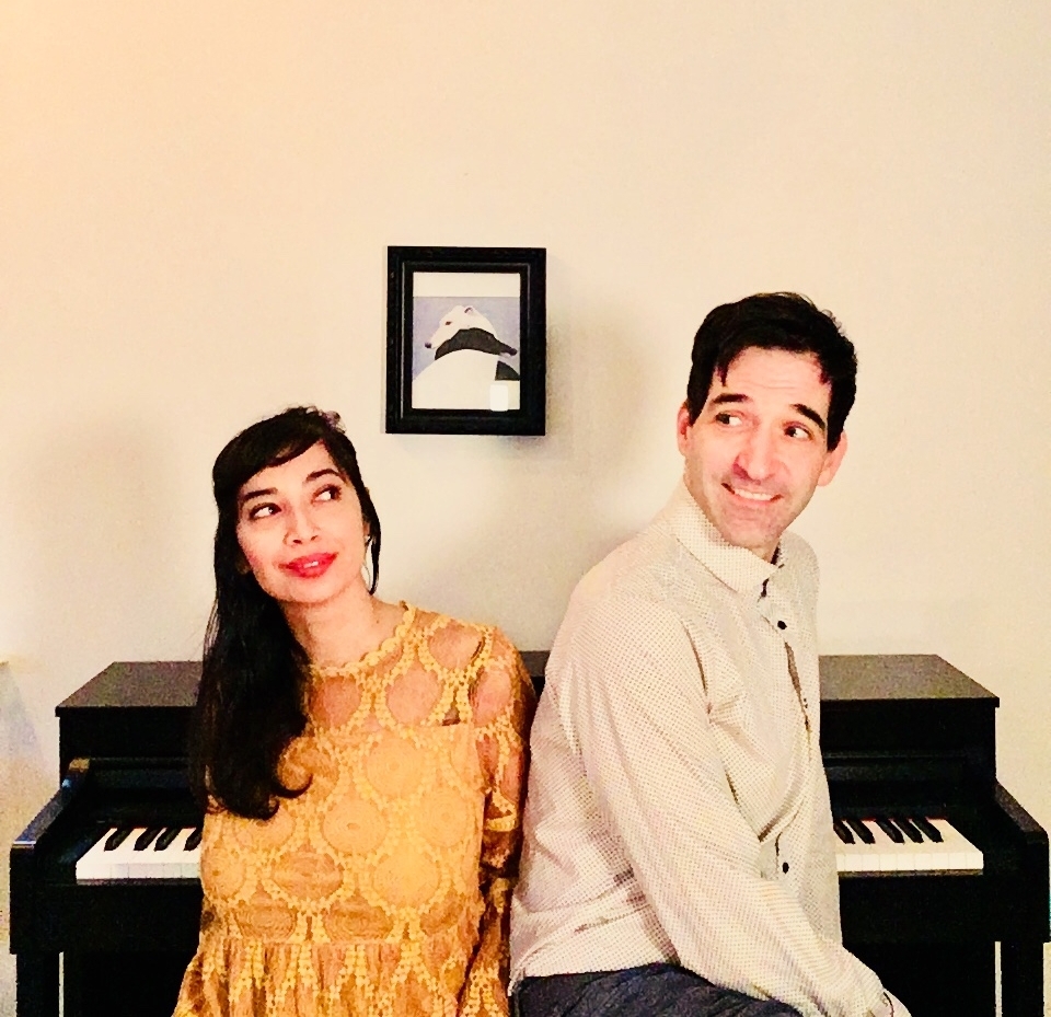 A photo of the main members of Mama Train: Sumitra and Philip, sitting in front of a piano. Sumitra wears a yellow dress and Philip a cream button down.
