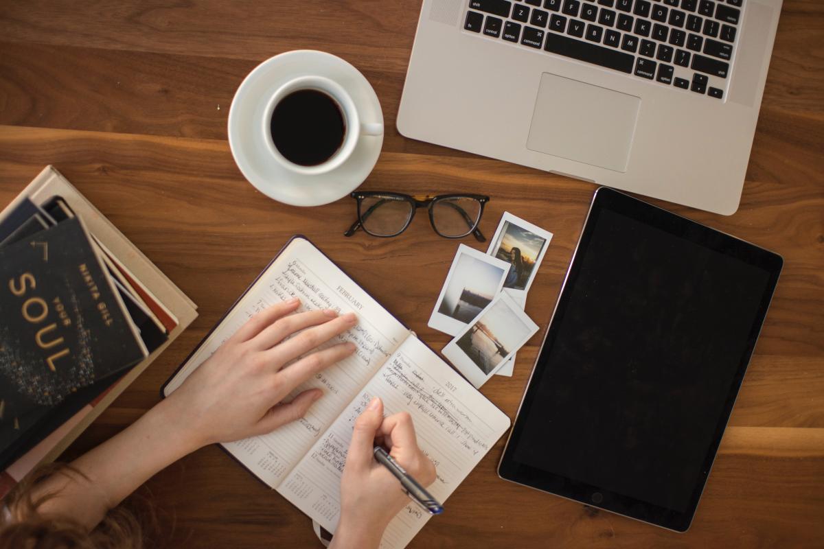 Photo of someone writing in a journal with computer, coffee, and glasses nearby. Photo by Thought Catalog on Unsplash